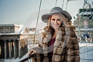 Portrait of happy brunette woman wearing re ress, fur mink winter coat and grey hat on the background of city