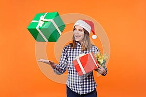 Portrait of happy brunette young woman in santa hat and checkered shirt standing throwing gift boxes in air or catching presents,