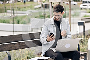 Portrait of happy business man enjoy success on mobile phone near modern office. Joyful guy reading good news on