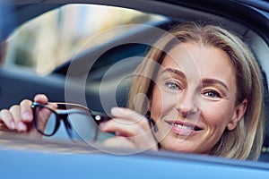 Portrait of a happy business woman holding eyeglasses and smiling at camera while sitting behind steering wheel of her