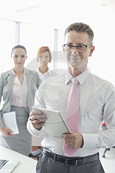 Portrait of happy businessman holding digital tablet with female colleagues in background at office