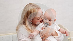 Portrait of happy caring smiling mother holding in her arms, hugging infant with pacifier, kissing, taking him out of