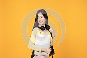 Portrait of happy casual Asian girl student with backpack, headphones and laptop isolated on yellow background. Back to school and
