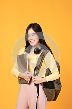 Portrait of happy casual Asian girl student with backpack, headphones and laptop isolated on yellow background. Back to school and