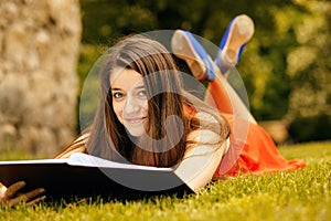 Portrait of happy casual woman in dress lying on green grass. She is reading a book