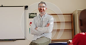 Portrait of happy caucasian male teacher sitting on desk in classroom looking at camera