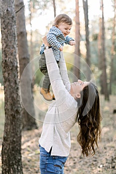 Portrait of happy Caucasian mother lifting and playing with her cute little son, enjoying their joint walk in pine