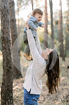 Portrait of happy Caucasian mother lifting and playing with her cute little son, enjoying their joint walk in pine