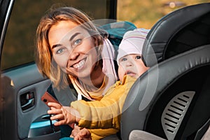 Portrait of happy caucasian mother prepares her little cute child for a trip by car. A woman fastens her seat belts in a