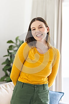 Portrait of happy caucasian woman standing with hands behind back in living room at home, copy space