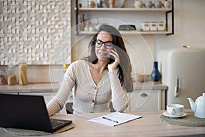 Portrait of happy caucasian woman working from home, talking on cellphone and using laptop computer, sitting in kitchen