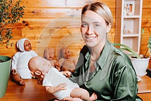 Portrait of happy caucasian young woman holding realistic newborn baby reborn doll in her arms and looking at camera, indoors