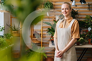 Portrait of happy charming female florist wearing apron standing posing in floral shop, looking at camera.
