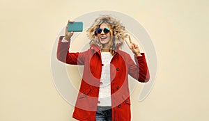 Portrait of happy cheerful laughing young woman taking selfie with smartphone having fun wearing red jacket coat with fur hood