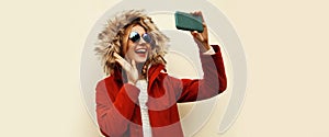 Portrait of happy cheerful laughing young woman taking selfie with smartphone having fun wearing red jacket with fur hood