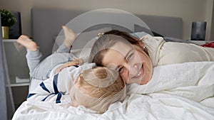 Portrait of happy cheerful mother lying in bed with her cute toddler son. Concept of family happiness, relaxing at home, having