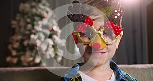 Portrait happy Child in festive Christmas glasses . Beautiful portrait smiling littlen girl sitting at home on the sofa