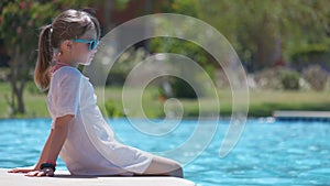 Portrait of happy child girl in white dress relaxing on swimming pool side on sunny summer day during tropical holidays