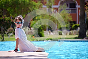 Portrait of happy child girl in white dress relaxing on swimming pool side on sunny summer day during tropical holidays