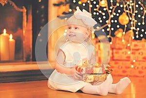 Portrait happy child little girl sitting with gift box looking away near christmas tree with garland lights and fireplace at