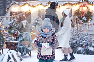 Portrait of a happy child standing on the evening street and holding out hands with gift box at snowy winter day. Christmas gifts