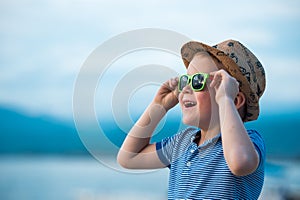 Portrait of happy child in sunglasses on the beach. Summer vacation concept. Smiling kid boy in hat and glasses near sea