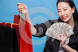 Portrait of a happy Chinese woman in black shirt holding red shopping bag and money isolated on a blue background