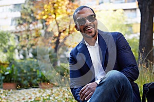 Portrait of Happy confident young African-American businessman in formal wear and sunglasses sitting at city park