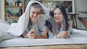 Portrait of happy couple lying in bed under blanket looking at camera laughing and smiling. Loving married people and