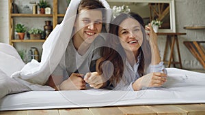 Portrait of happy couple lying in bed under blanket then showing faces looking at camera laughing and smiling. Loving