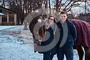 Portrait of a happy couple near to a brown horse on the ranch on a winter day.