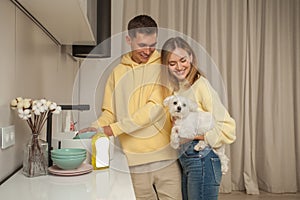 Portrait of happy couple in yellow hoodies, man washing the dishes, woman holding little white dog