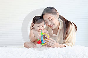 Portrait of happy crawling baby sits on fluffy white rug with mother, little cute kid girl plays with developmental toys, mom hugs