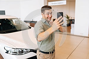 Portrait of happy customer male client in casual clothes doing selfie shot on mobile smartphone at dealership after