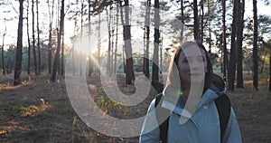 Portrait of happy cute good-looking female traveler in blue jacket with big tourist backpack walking through autumn forest at sunr