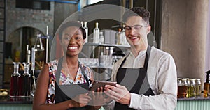 Portrait of happy diverse couple wearing aprons working at a bar, using tablet and smiling to camera