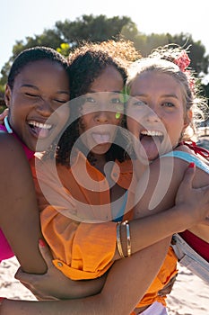 Portrait of happy diverse female friends embracing and sticking tongue out at beach, with copy space
