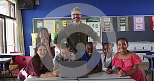Portrait of happy diverse male teacher and group of schoolchildren looking at laptop
