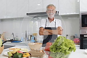 Portrait of happy elderly man with apron standing with arms crosse at kitchen with colorful fresh vegetables, fruits,ingredients.