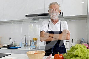Portrait of happy elderly man with apron standing with arms crosse at kitchen with colorful fresh vegetables, fruits,ingredients.