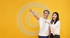 Portrait of happy excited Asian couple tourists pointing hands to empty space on isolated light yellow background