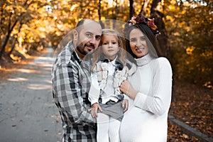 Portrait of happy family on background of golden park in autumn day. Father, daughter and pregnant mother