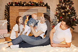 Portrait of happy family of father, mother and two daughters sitting on floor on soft carpet near fireplace and xmas tree,