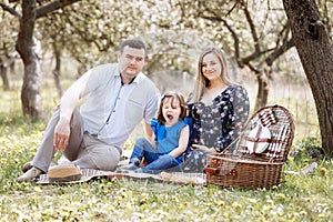 Portrait of happy family on picnic. pregnant Mom, dad and daughter sitting in spring blooming park. Young family spending time