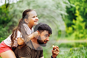 Portrait of happy family sitting on picnic in park forest around trees bushes. Little daughter sitting on fathers back.