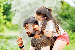 Portrait of happy family sitting on picnic in park forest around trees bushes. Little daughter sitting on fathers back.