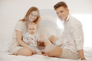 Portrait of happy family of three on white background. Mother, father and infant baby sit on bed and hug. Photo of young