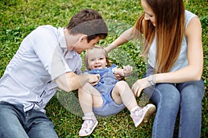 Portrait happy family of three. Young parents play with their little daughter sitting on the lawn in the Park.