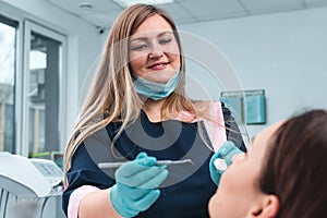 Portrait of happy female dentist in mask sitting, holding dentist tools, smiling and examining young woman in dental chair,