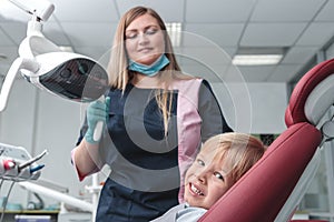 Portrait of happy female dentist in mask standing, holding dentist tools, smiling and examining young boy in dental chair, healthy